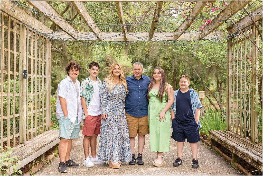 Family of 6 is standing in a open gazebo surrounded by lush greenery. Son is wearing white shirt and green shorts. Second son is wearing  a green and coral shirt over a white shirt and coral shorts. Mom is wearing a long sleeve ankle length blue dress with blue print. Dad is wearing a button up blue shirt and brown pants. Daughter is wearing a lime green sleeveless dress. 3rd son is wearing a black shirt and black shorts with a short sleeve blue shirt over the black shirt. 
