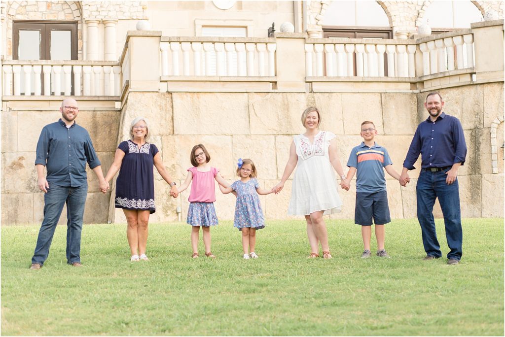 Family of 7 are standing just in front of large stone court house. Dad is wearing dark blue long sleeve button up shirt with jeans. Grandma is wearing a navy blue short sleeve dress with white trim. 1 daughter is wearing a pink shirt and blue skirt. 2nd daughter is wearing a short sleeve blue dress with blue and pink print. Mom is wearing a short sleeve knee length white dress. Son is wearing a blue short sleeve polo with a large orange stripe and small white stripe. Dad is wearing a long sleeve button up navy shirt and jeans. 