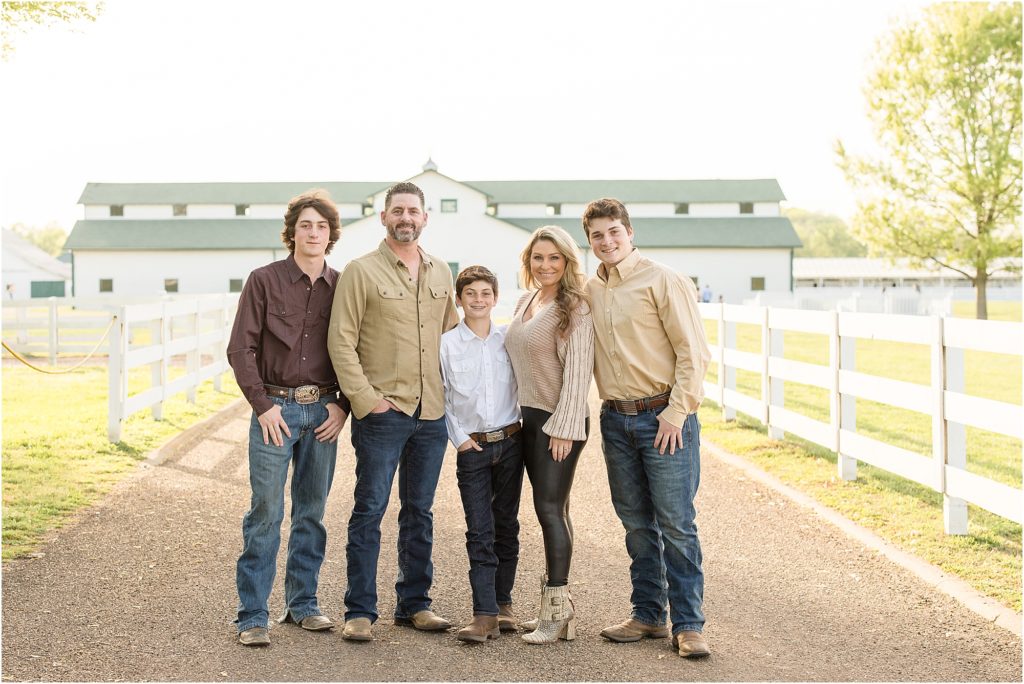 Beautiful family of 5 are standing with a huge white and green barn in the background and in between white wooden fence. 1st son is wearing a long sleeve button up brown shirt and blue jeans. Dad is wearing a light brown long sleeve button up shirt and blue jeans. 2nd son is a wearing a long sleeve button up white shirt and blue jeans. Mom is wearing long sleeve gold sweater and black leggings and gold booties. 3rd son is waring a long sleeve button up light brown shirt and blue jeans. 