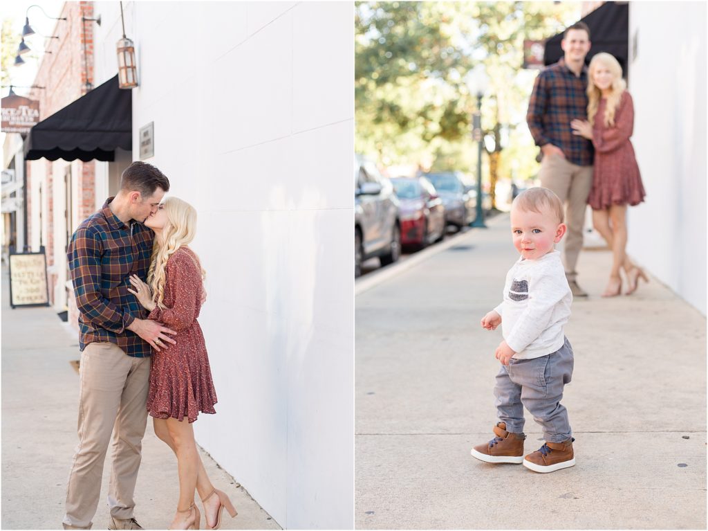A beautiful family of 3 is standing on the sidewalk. Dad is wearing a brown and blue plaid shirt and tan pants. Mom is wearing a long sleeve dark pink short dress. Baby is wearing a long sleeve white shirt with a grey pocket and grey pants.