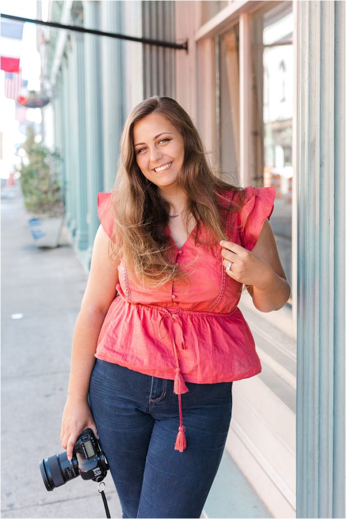 Kelsey is wearing a pink flutter sleeve shirt with a tie at the waist with tassels on the end. She is wearing blue jeans and holding a camera.