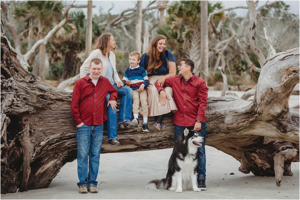 Family of 5 standing on the beach. Kelsey is wearing a long taupe pleated skirt and a denim flutter sleeve shirt. Her mom is wearing blue jeans, a blue shirt, and a cream cardigan. Brother is wearing a red long sleeve button-up shirt with blue jeans. The little boy is wearing a blue white and red striped long-sleeve shirt and khaki pants. Seated on the beach as well is a black and white dog. 