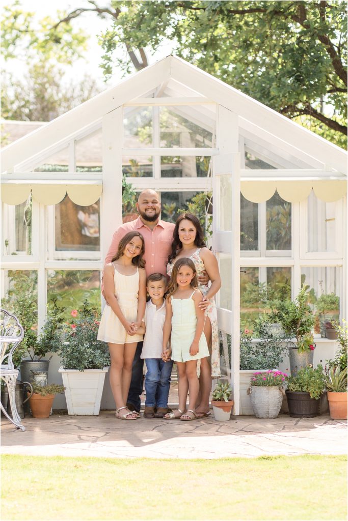 A family of 5 is standing in front of a white greenhouse with large windows. Mom is wearing a white tank and jean shorts. Sitting in her lap is her son who is wearing a short sleeve polo and blue jeans. Dad is wearing a short sleeve green shirt and blue jeans. His daughter is wearing a mint-colored short sleeveless romper. My oldest sister is wearing a short sleeveless pale yellow romper. 