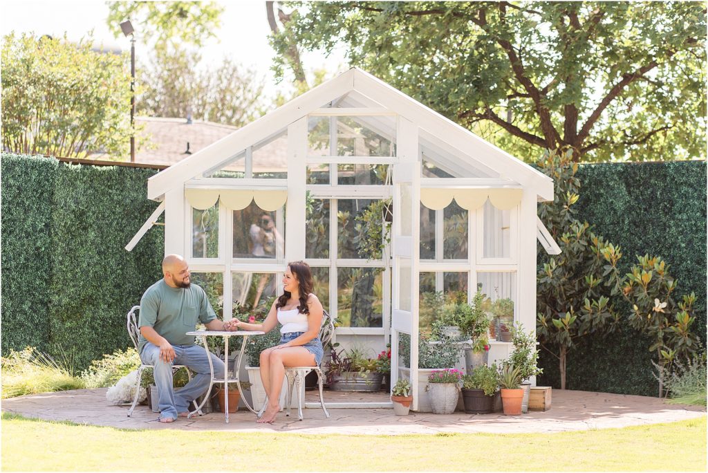 Husband and wife are sitting just outside the greenhouse at a white rod iron table and chairs. The husband is wearing a short sleeve green shirt and blue jeans. The wife is wearing a white tank and blue jeans. 