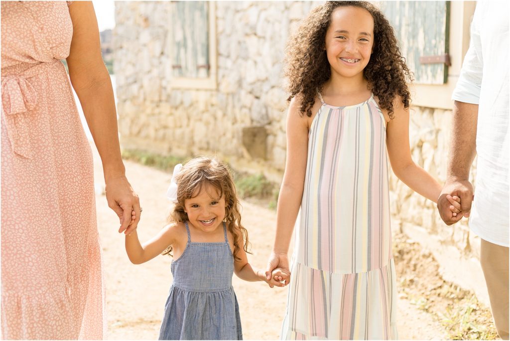 Sisters are walking with a stone wall in the background and green shutters. 1st sister is wearing a sleeveless long pastel striped dress. Little sister is wearing a sleeveless blue dress.