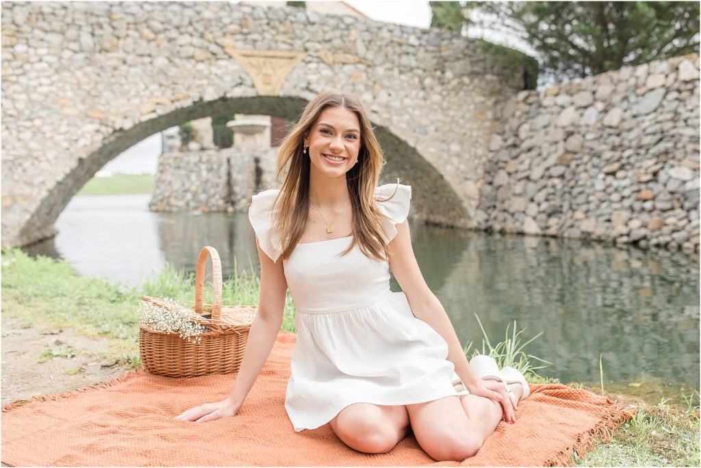 The young lady sitting on an orange blanket and picnic basket. She is wearing a white flutter sleeve dress and white shoes. With a beautiful stone bridge and wall in the background.