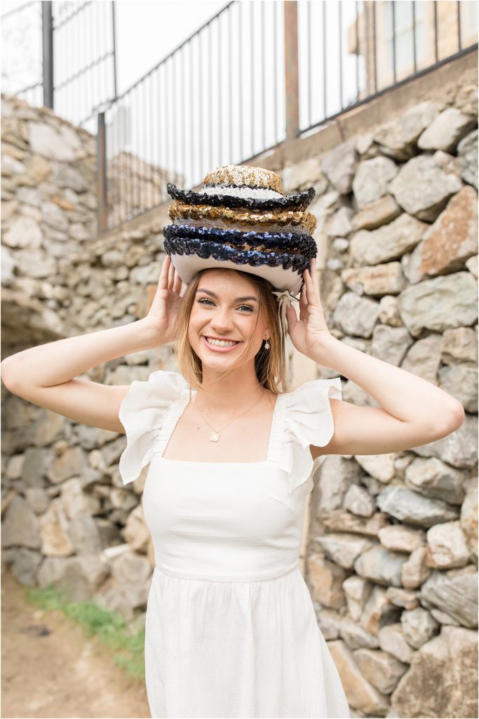 The young lady is standing in front of a stone wall wearing a white flutter sleeve dress. She is wearing several drill team hats,