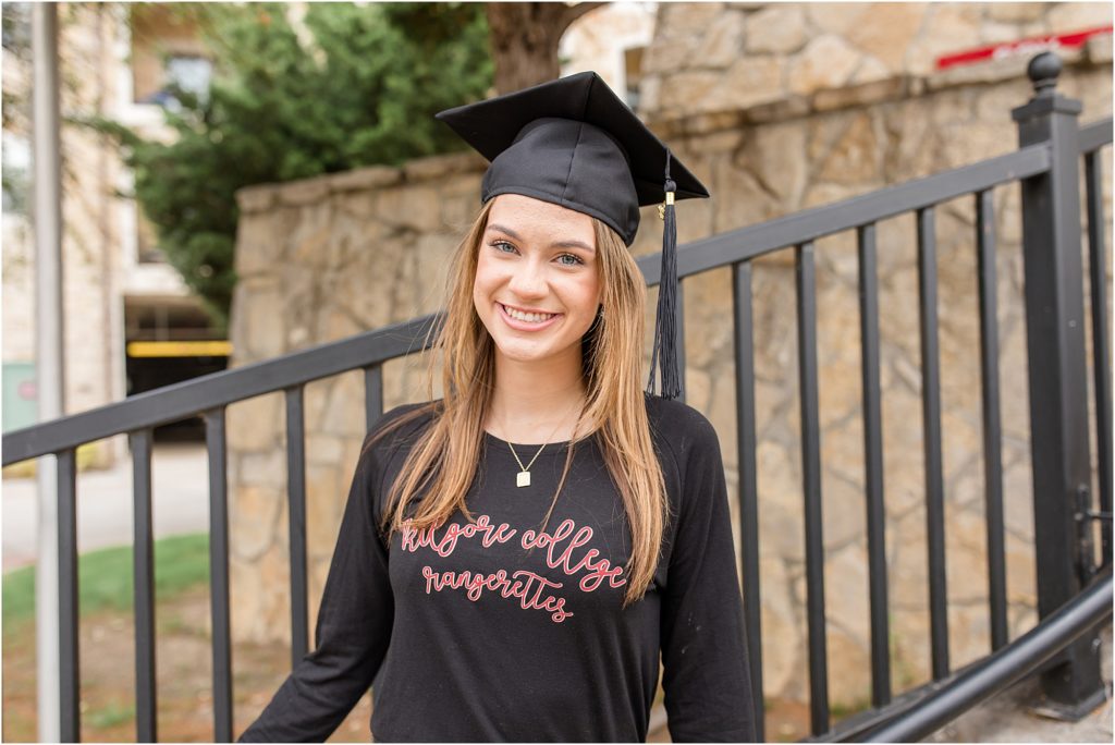 The young lady is standing just in front of a stone wall and black metal handrail. She is wearing her graduation cap and a long sleeve black shirt.