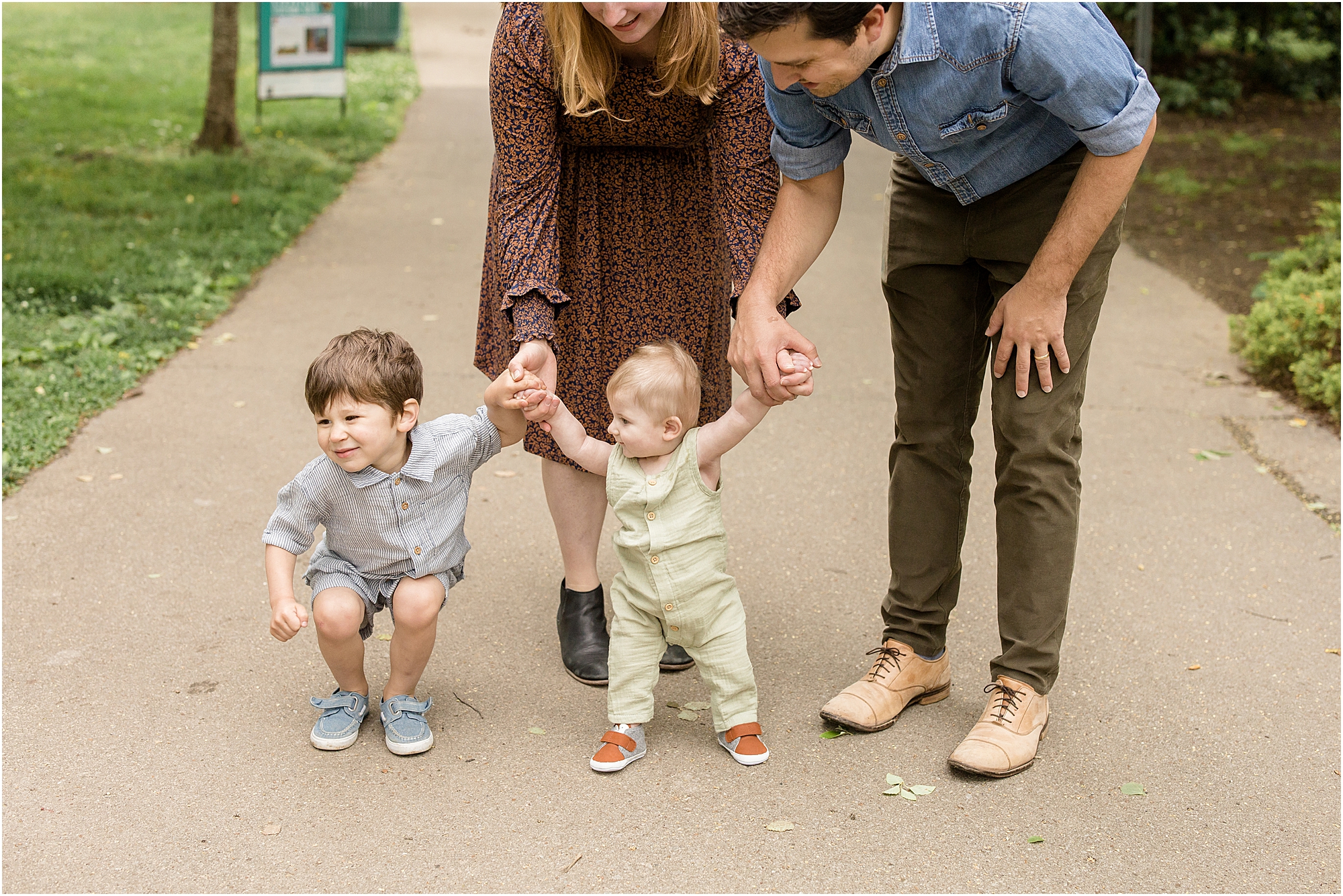Family of 4 standing on sidewalk holding hands. Mom is wearing a long sleeve black and orange dress. Dad is wearing a long sleeve blue jean shirt with the sleeves rolled and olive green pants. The boys are wearing a light blue short sleeve shirt and shorts and blue velcro shoes and the other is wearing a light green sleeveless long romper with orange shoes