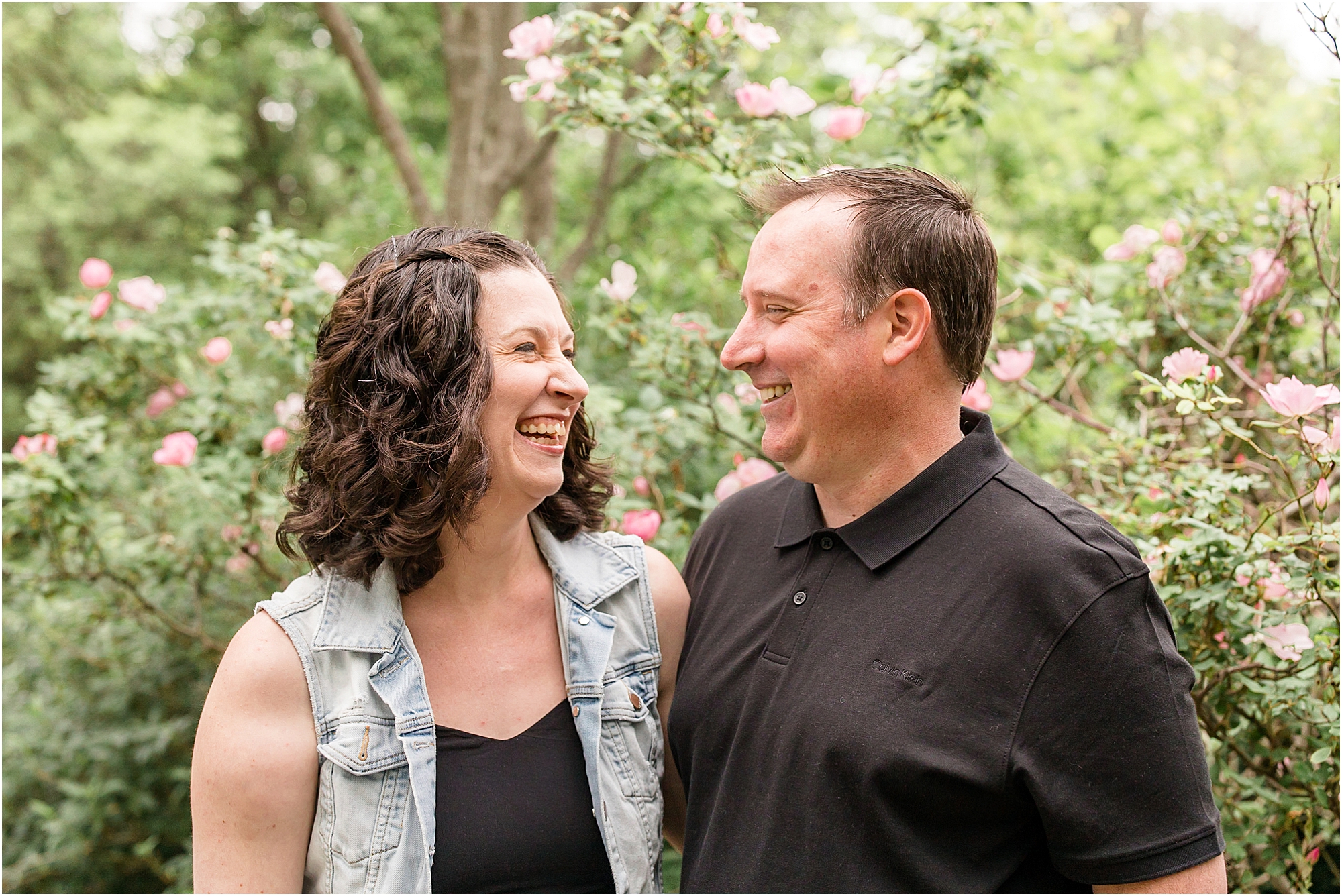 Husband and wife are standing in front of beautiful flowering trees. Mom is wearing a sleeveless black dress and a denim vest. Dad is wearing a black polo.