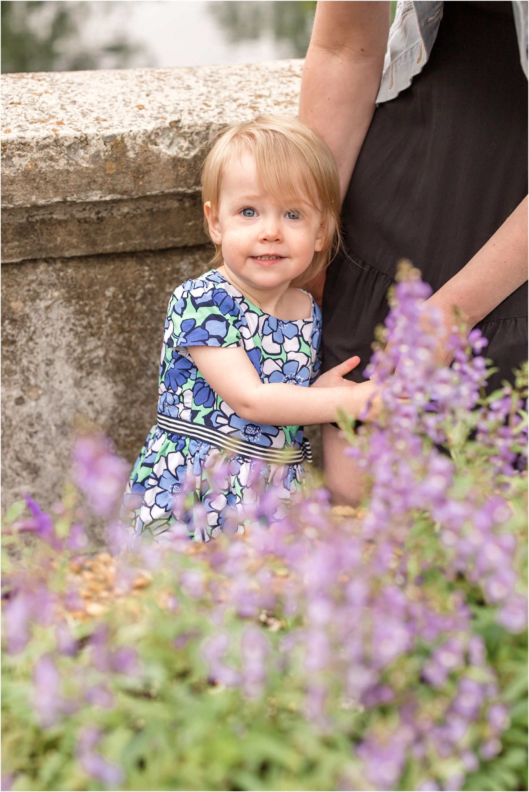 The little girl is standing in front of a stone wall, behind a flowering bush. She is wearing a white, blue, and green floral print dress.
