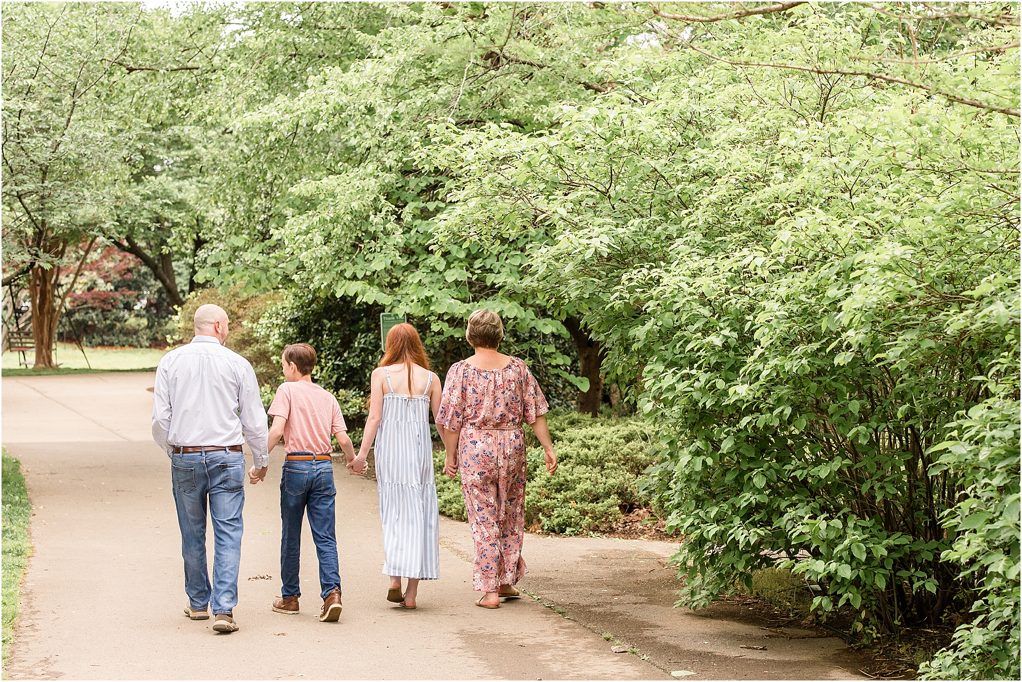 A family of 4 is walking down the sidewalk surrounded by beautiful greenery. Mom is wearing a pink floral jumpsuit. The daughter is wearing a sleeveless blue and white dress. Son is wearing a pink shirt and blue jeans. Dad is wearing a long sleeve button white shirt and blue jeans.