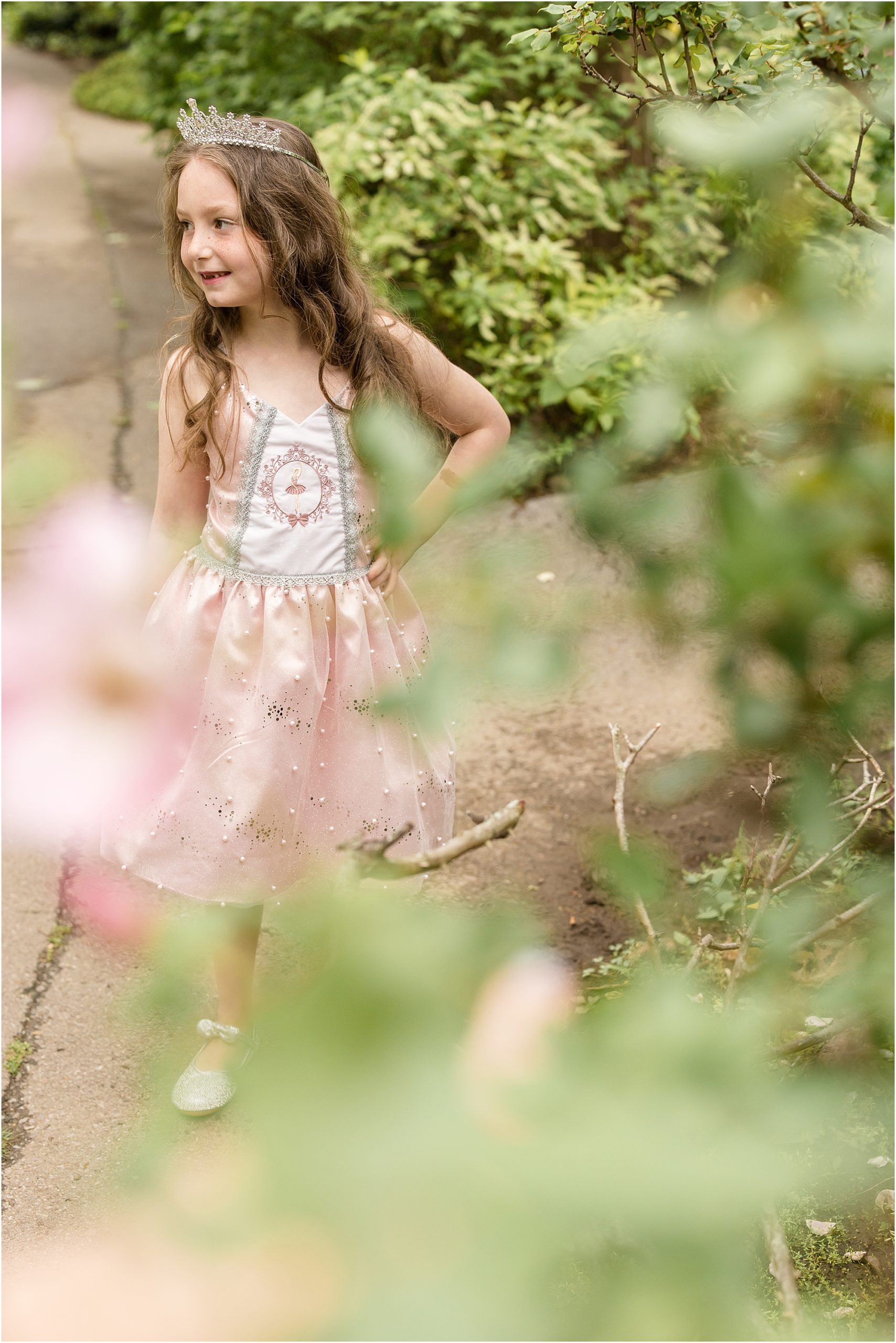 The little girl is standing surrounded by greenery. She is wearing a pink princess dress and a tiara. 