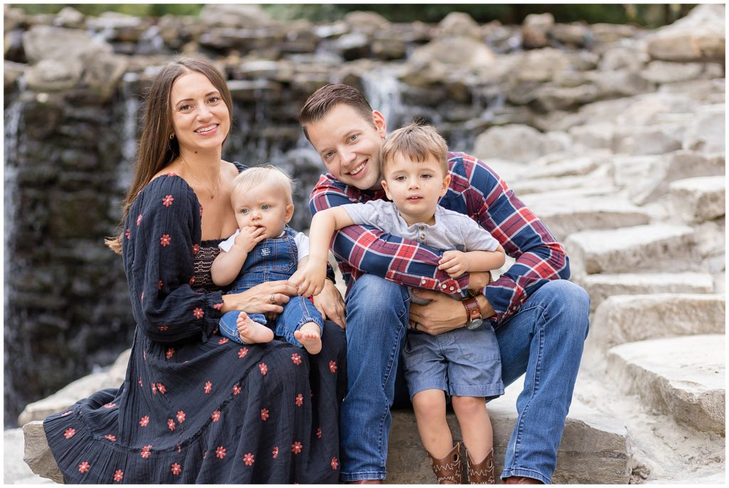 Family of 4 sit on rocks in front of waterfall while dad hugs son and mom holds baby in lap. We had such a wonderful time shooting this sweet family during their family session with Wisp + Willow Photography Co. 