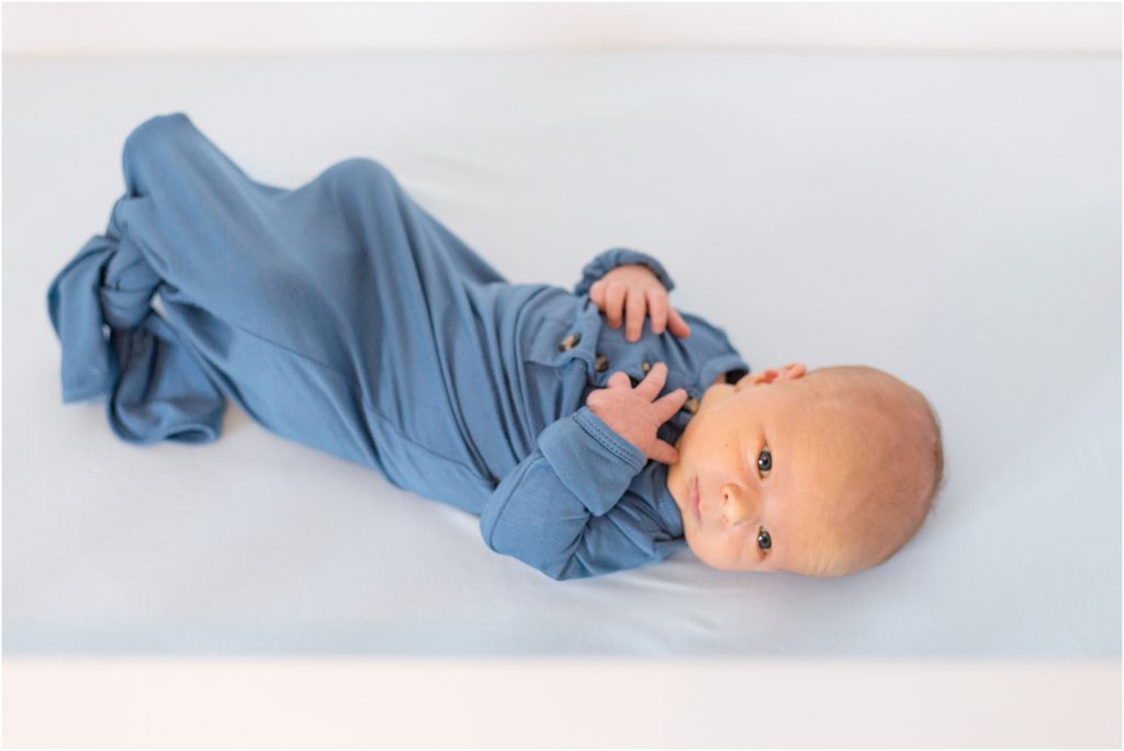 The baby boy is laying on the bed and wearing a blue sleep sack.
