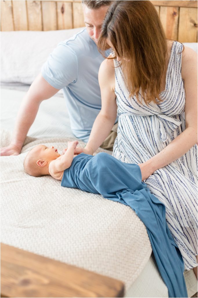 Cute family of 3 sitting on the bed. Mom is wearing a sleeveless gauze-striped dress. Dad is wearing a light blue shirt and khaki shorts. The baby boy is swaddled in blue.