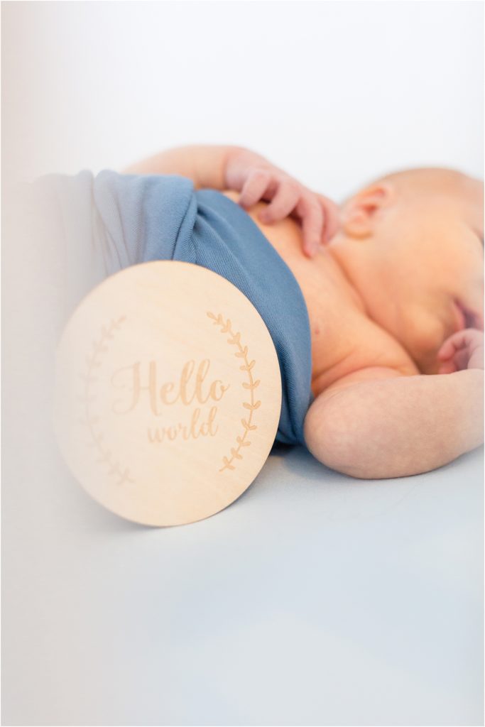 The baby boy is laying on the bed swaddled in blue with a wooden Hello World sign. 