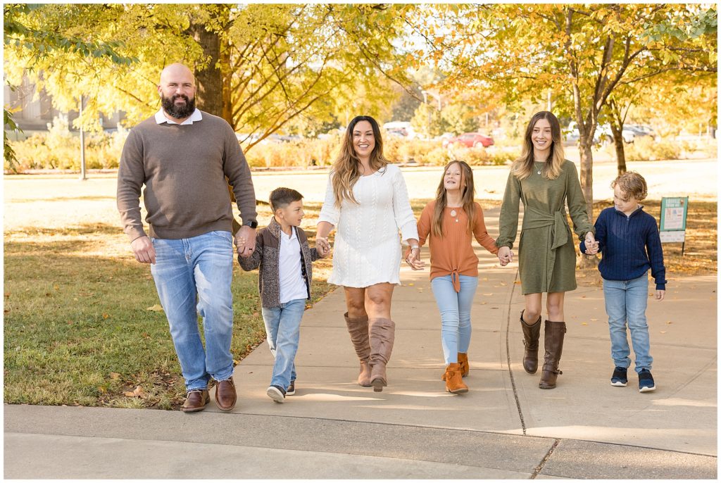 Family of 6 walk hand in hand down sidewalk with beautiful autumn trees behind them during fall family session. 