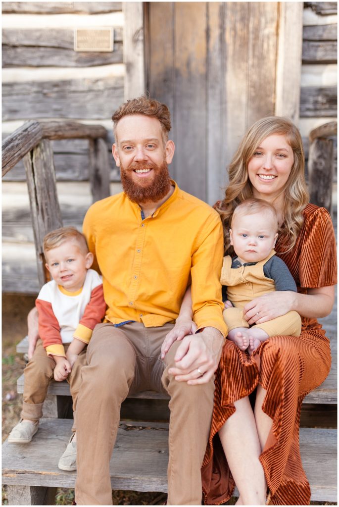 Mom and dad wearing yellow and orange hold 2 sons on lap while sitting on log home steps during fall family photo session with Wisp + Willow Photography Team. 