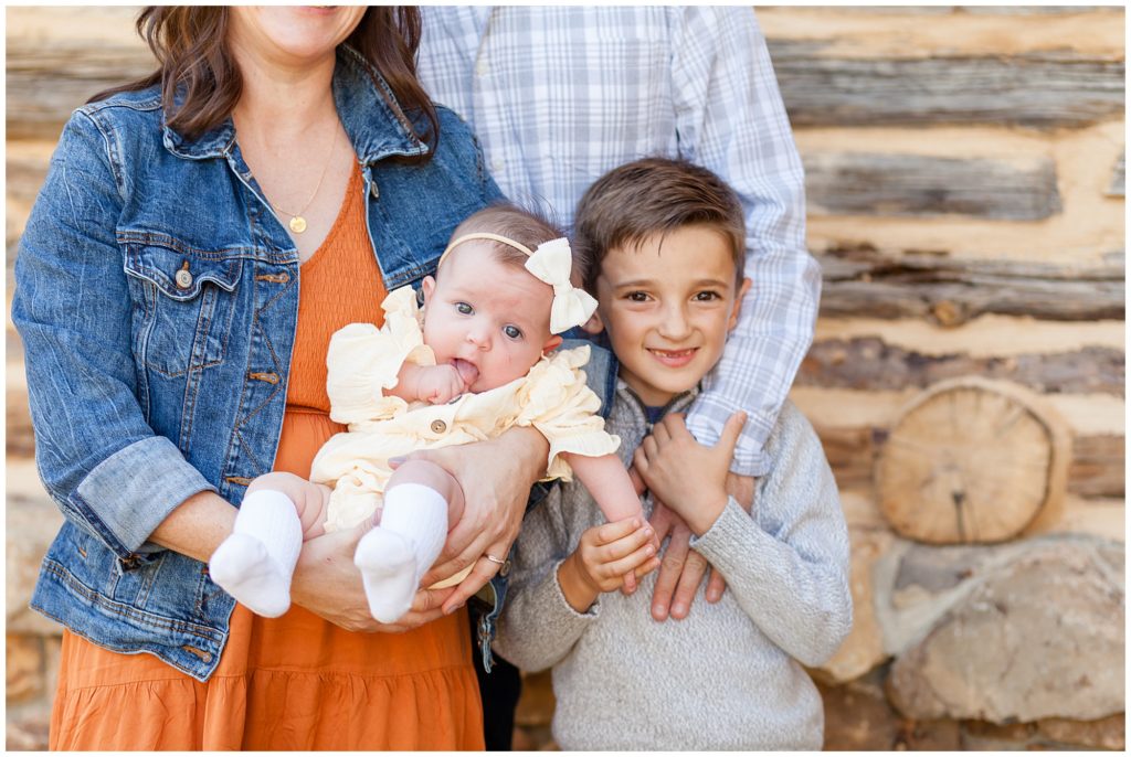 Mom in orange dress and jean jacket holds baby girl and dad in grey plaid button down has his hand on sons chest during fall family session. 