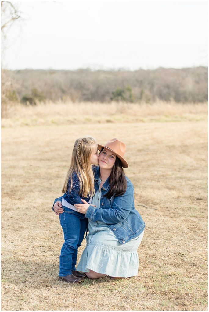 Daughter kisses mom in light blue dress and jean jacket on cheek in wide open field during family session with Wisp + Willow Family Photography. 
