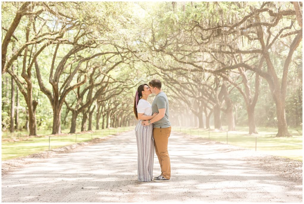 Couple kisses while standing under row of trees during family session with Wisp + Willow Photography Co. 