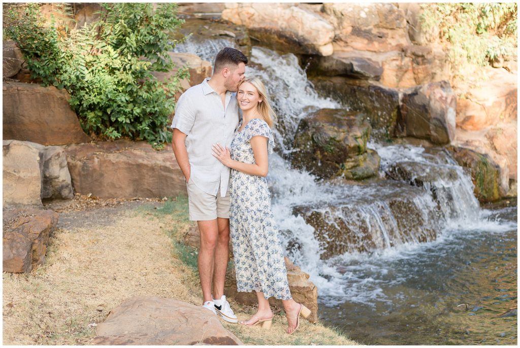 Man in grey button down kisses his gorgeous fiance in blue floral dress on forehead in front of waterfall. 