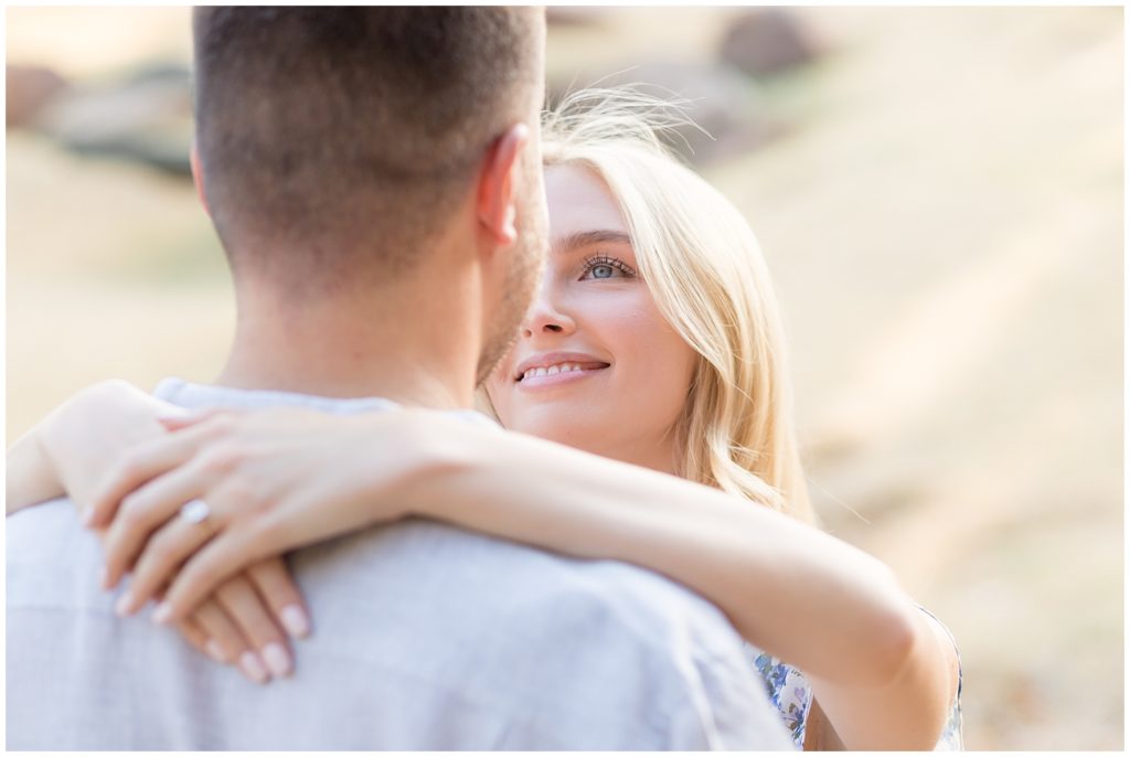 Beautiful woman with blonde hair gazes into fiance's eyes while wrapping her arms around his neck during engagement session. 