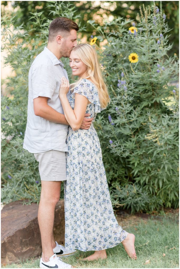 Woman in blue floral dress places her hand on fiance's chest while he kisses her on forehead in front of flower bush in the summer time during engagement photo session. 