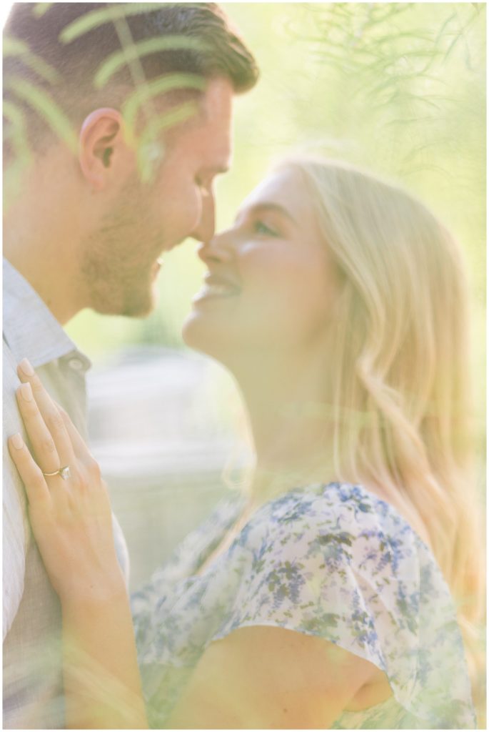 Engaged man and woman smile as they lean in to kiss while she places her hand on his chest in the sunlight. 