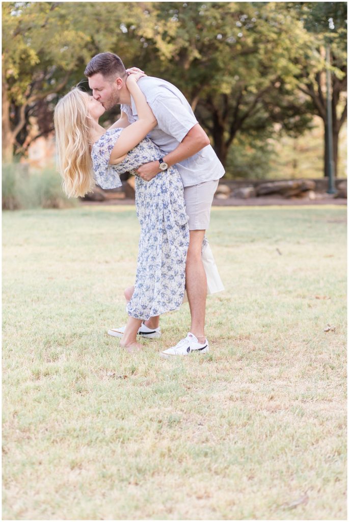 Man in grey button down leans over to kiss his beautiful blonde fiance in a blue floral dress while standing in the grass in the summer time during engagement shoot. 