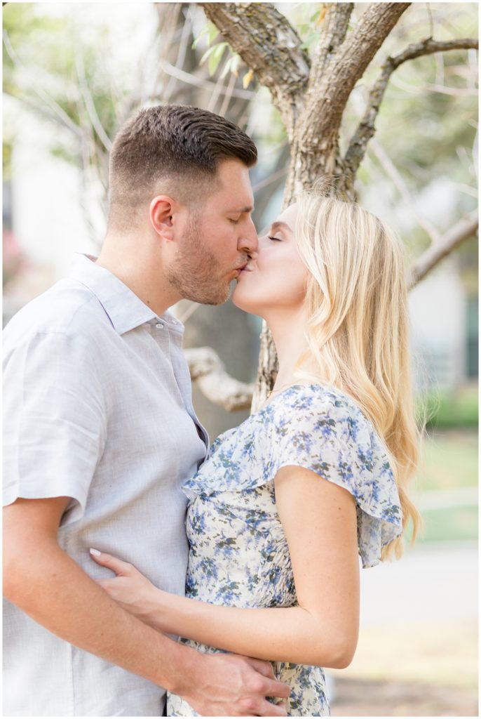 Engaged man and woman hold each other and kiss in front of tree outside in Frisco Central Park during photo session. 