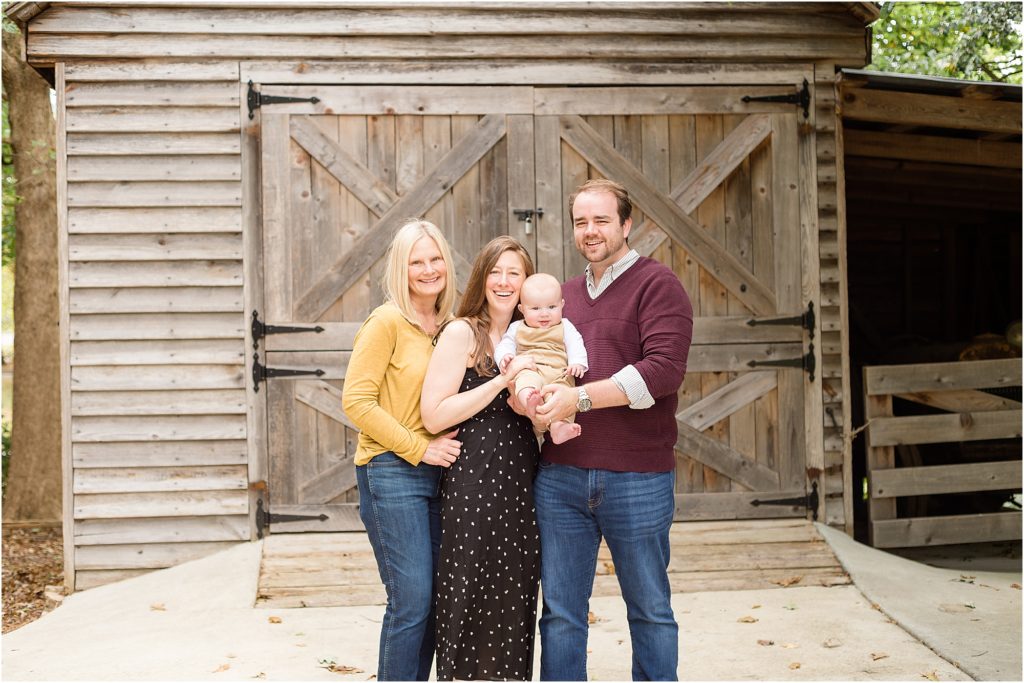 A family of 4 is standing just in front of a rustic wooded barn. Mom is wearing a sleeveless black dress. Dad is wearing a burgundy sweater. Grandma si wearing a long sleeve mustard yellow shirt and blue jeans and the baby is wearing tan overalls and a long shelve white shirt.