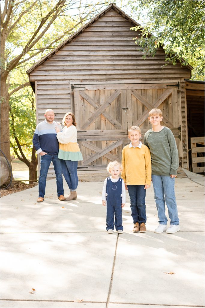 The family of 5 is standing with the wooden board and fence in the background. Mom is wearing a cream, yellow and green tiered shirt and blue jeans. Son is wearing long sleeve green shirt and blue jeans, the son is wearing a long navy overall romper and white shirt. 3rd son is wearing a mustard yellow long-sleeve sweater and blue jeans. Dad is wearing 2 shades of blue long-sleeve sweaters. 