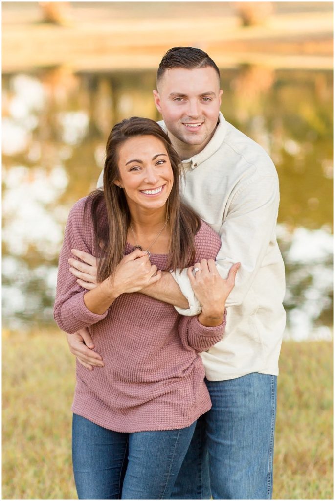 Couple in mauve and cream shirts hug and smile at the camera in front of lake. 