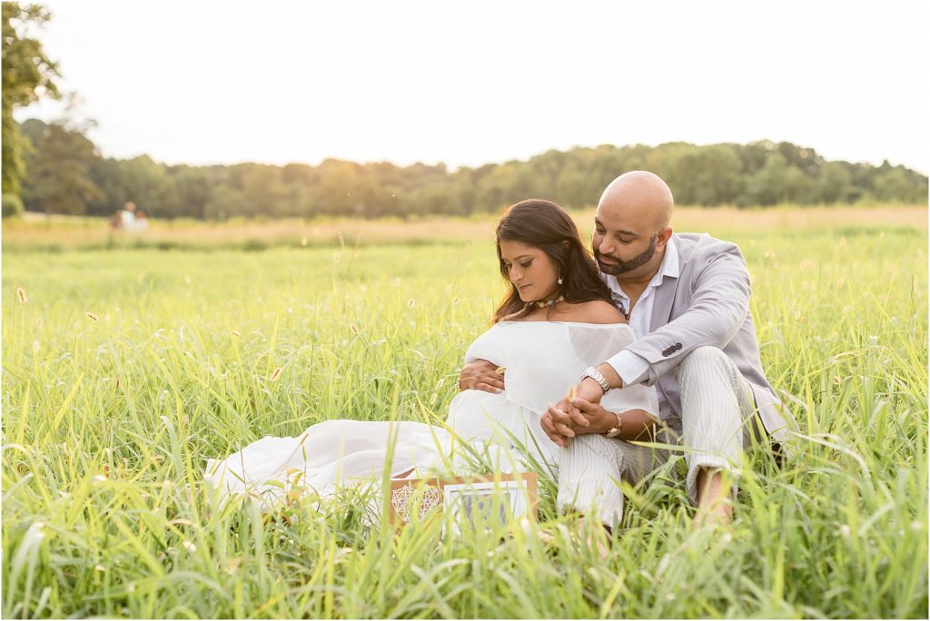 This beautiful parents-to-be are seated in a grassy field. Mom-to-be is wearing an off-the-shoulder white dress. Dad is wearing a grey suit.