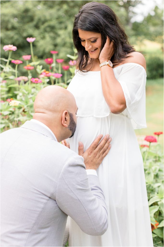 This beautiful parents-to-be are standing just in front of pink flowers. Dad-to-be is kissing mommy's tummy. He is wearing a grey suit. Mom-to-be is wearing a long off-the-shoulder dress.