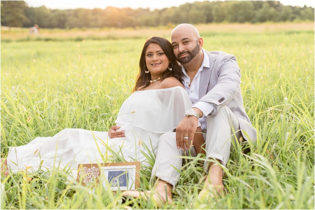 Parents-to-be are seated in a large open grass field with a sweet sonogram in a frame. Mom is wearing a long off-the-shoulder white dress. Dad is wearing a grey suit