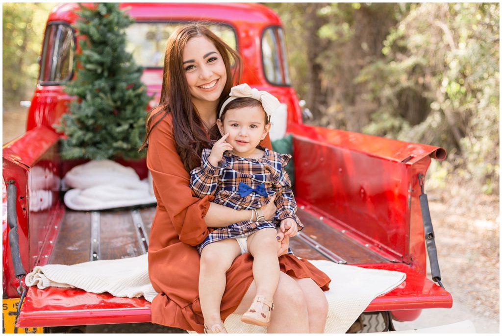 Daughter sits in mom's lap in bed of red pickup truck with tree in the back during Christmas photo session. 