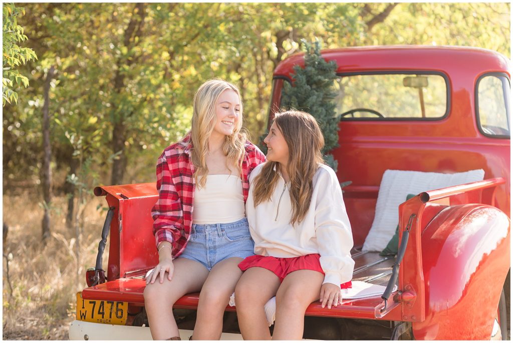 2 sisters sit in bed of red pickup truck while smiling at each other during Christmas photo session.