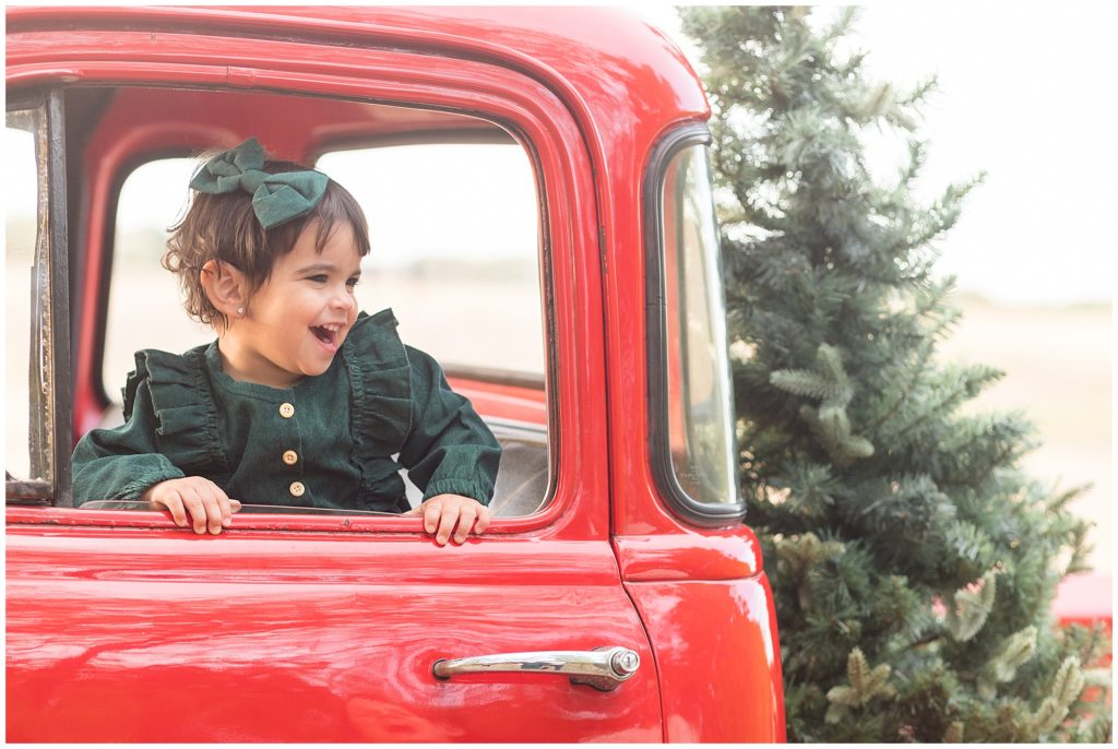 Baby girl in green dress and bow laughs as she looks out window of red pickup truck during Christmas family session. 