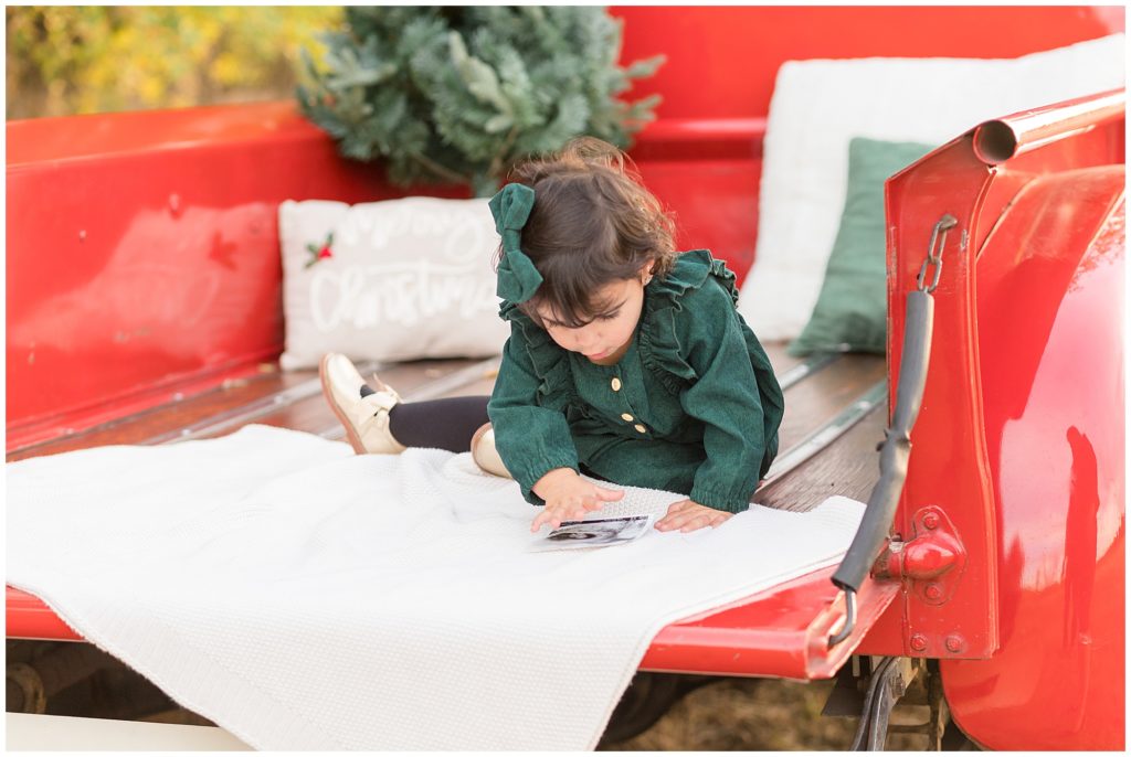 Baby girl in green dress plays in back of red pickup truck on cream blanket with tree in the back during Christmas photo session. 
