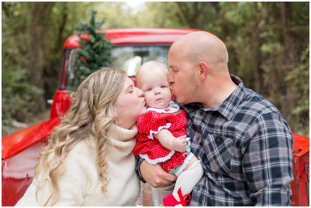 Mom and dad kiss baby girl in red dress while sitting in bed of red pickup truck during Christmas family session. 