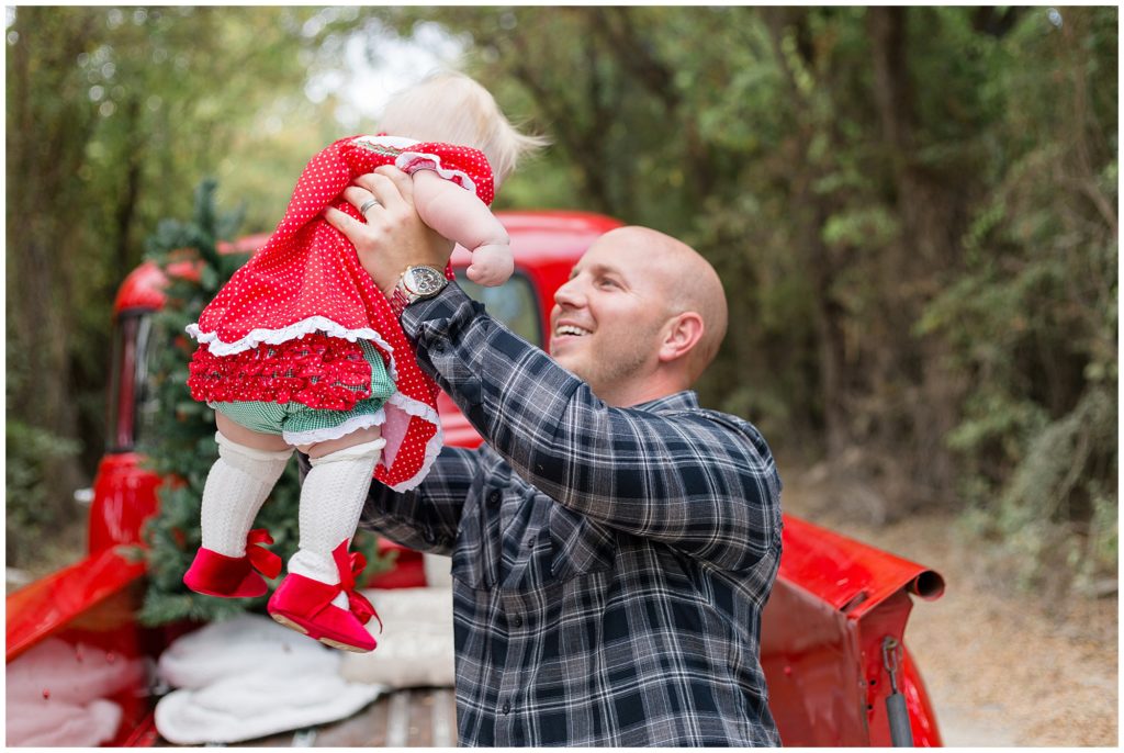Dad in grey plaid shirt holds up baby girl in red and green Christmas dress and shoes in front of red pickup truck.
