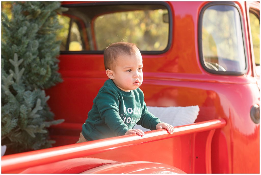 Baby boy in green Christmas sweatshirt stands in bed of red pickup truck with tree in back in Frisco, TX. 
