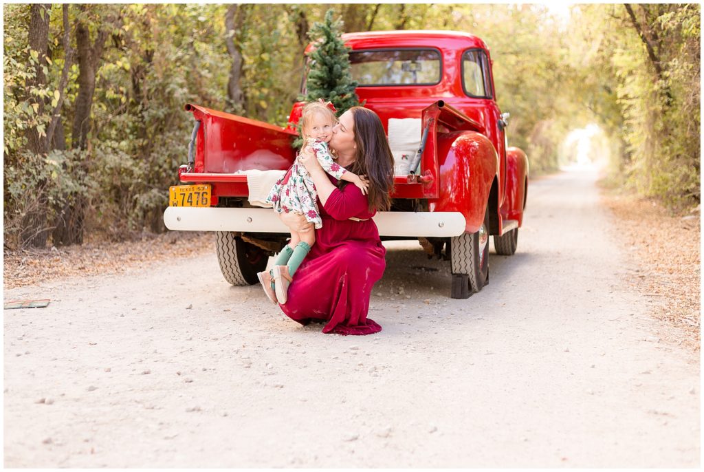 Mom in red dress kisses daughter while in front of red pickup truck with tree in back during Christmas photos in Frisco, TX. 