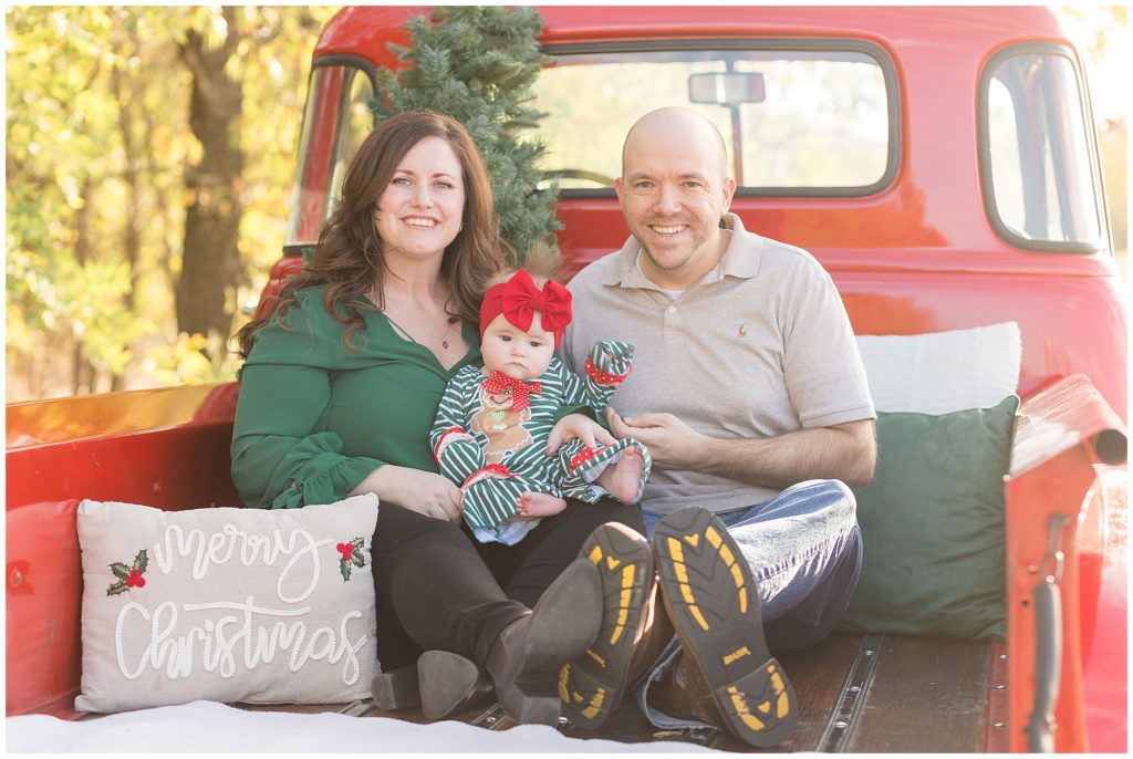 Mom and dad sit in bed of red pickup truck while holding baby with red bow during Christmas family session in Frisco, TX. 