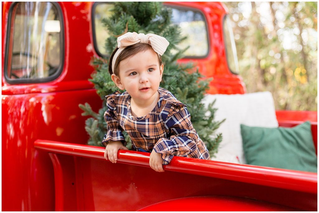 Little girl stands in bed of red pickup truck in plaid dress and cream bow with Christmas tree behind her during Christmas photo session in Frisco, TX. 