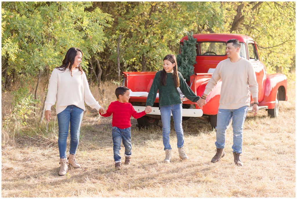 Family of 4 hold hands in front of red pickup truck with tree in back during Christmas photo session in Frisco, TX. 