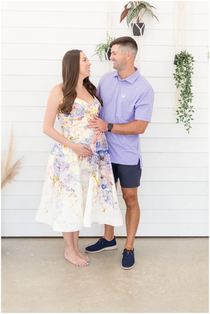 Husband in purple polo and wife in purple floral dress smile at each other during studio maternity shoot. 